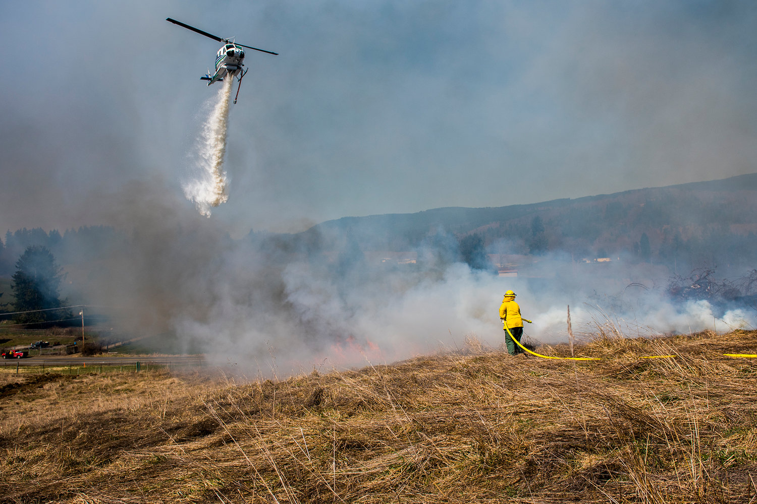 FILE PHOT0 — A helicopter used by the Wildfire Aviation Division of the Department of Natural Resources drops water from the sky onto a hillside while a DNR firefighter uses a hose to extinguish flames in Mossyrock in March of 2019.