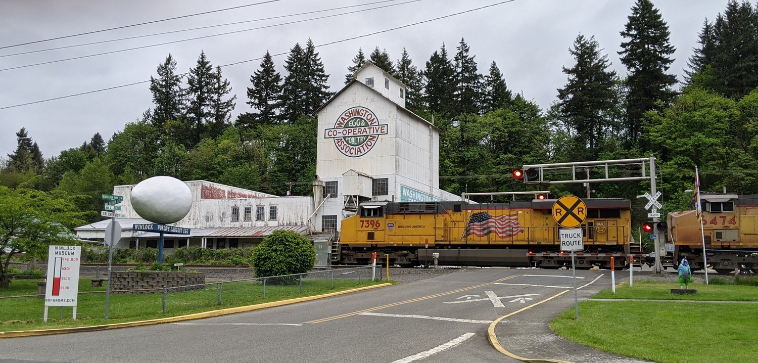 A train races past the historic Winlock hatchery and renowned giant egg in the city of Winlock. In the foreground is the painted wooden thermometer sign showing current fundraising efforts to purchase a rare Winlock history collection before it’s too late, for the Winlock Historical Museum.
