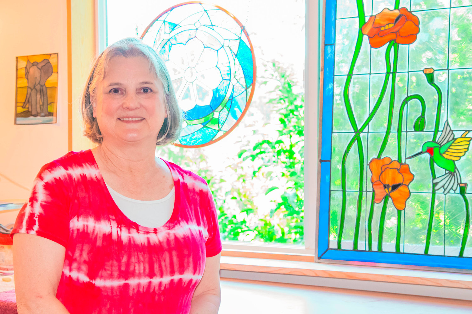 Marcy Anholt smiles and poses for a photo in front of stained glass in her studio.