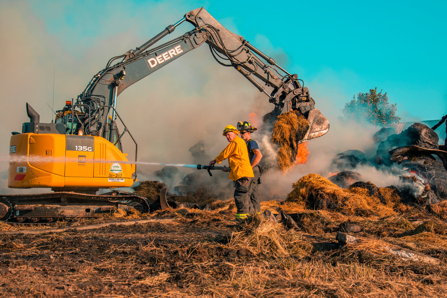 Firefighters use hoses to extinguish hot spots as an excavator lifts flaming bales of hay from remnants of a structure following a fire in Napavine.