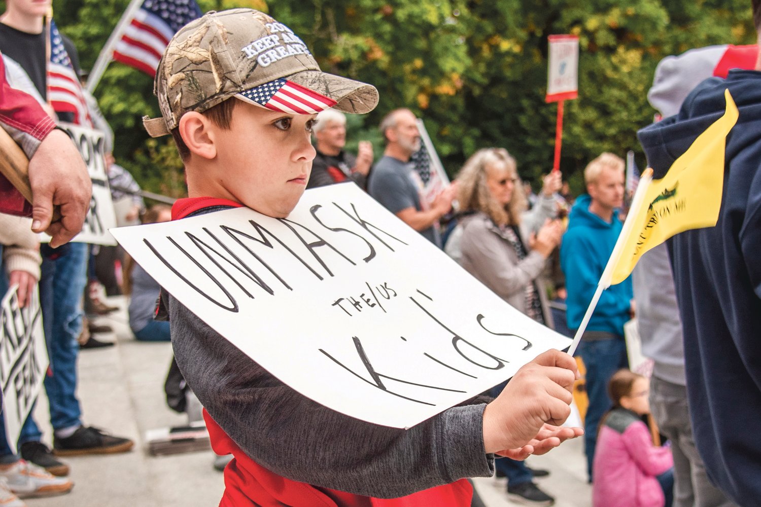 A young rally goer waves a flag while holding a sign that reads “Unmask the/us kids,” during a “Medical Freedom Rally” Sunday afternoon in Olympia.