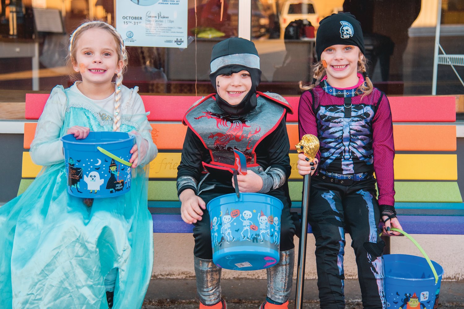 From left, Lillian dressed as Elsa, David dressed as a Dragon Ninja, and Anika dressed as a Dark Rebel smile and pose for a photo while sitting on a Buddy Bench in Morton on Halloween.