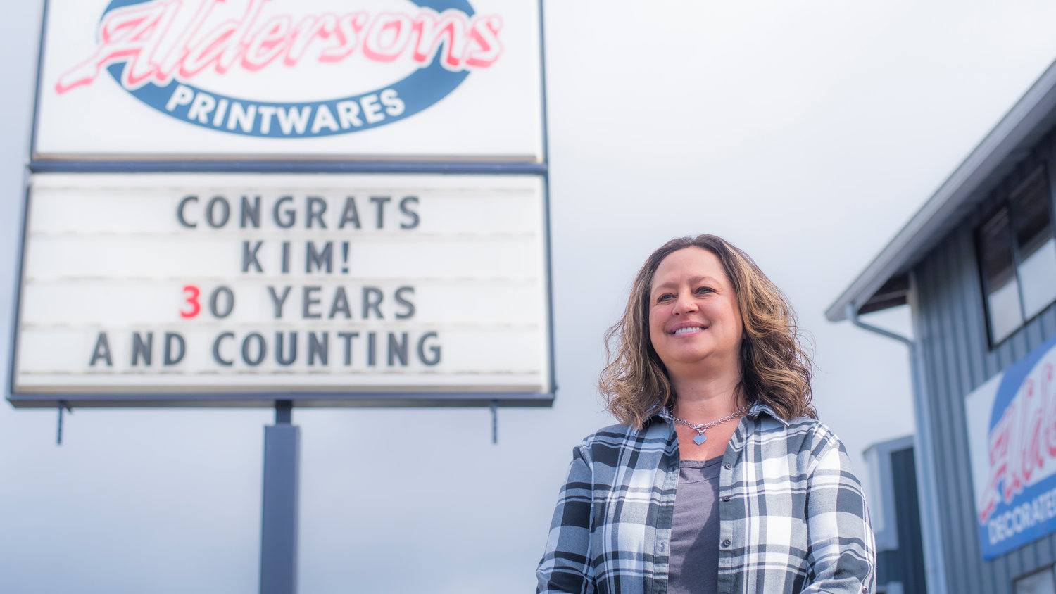 Kim Schrader smiles in front of a sign congratulating her for 30 years of employment on Tuesday in Centralia outside of Alderson’s Awards West-Printwares.