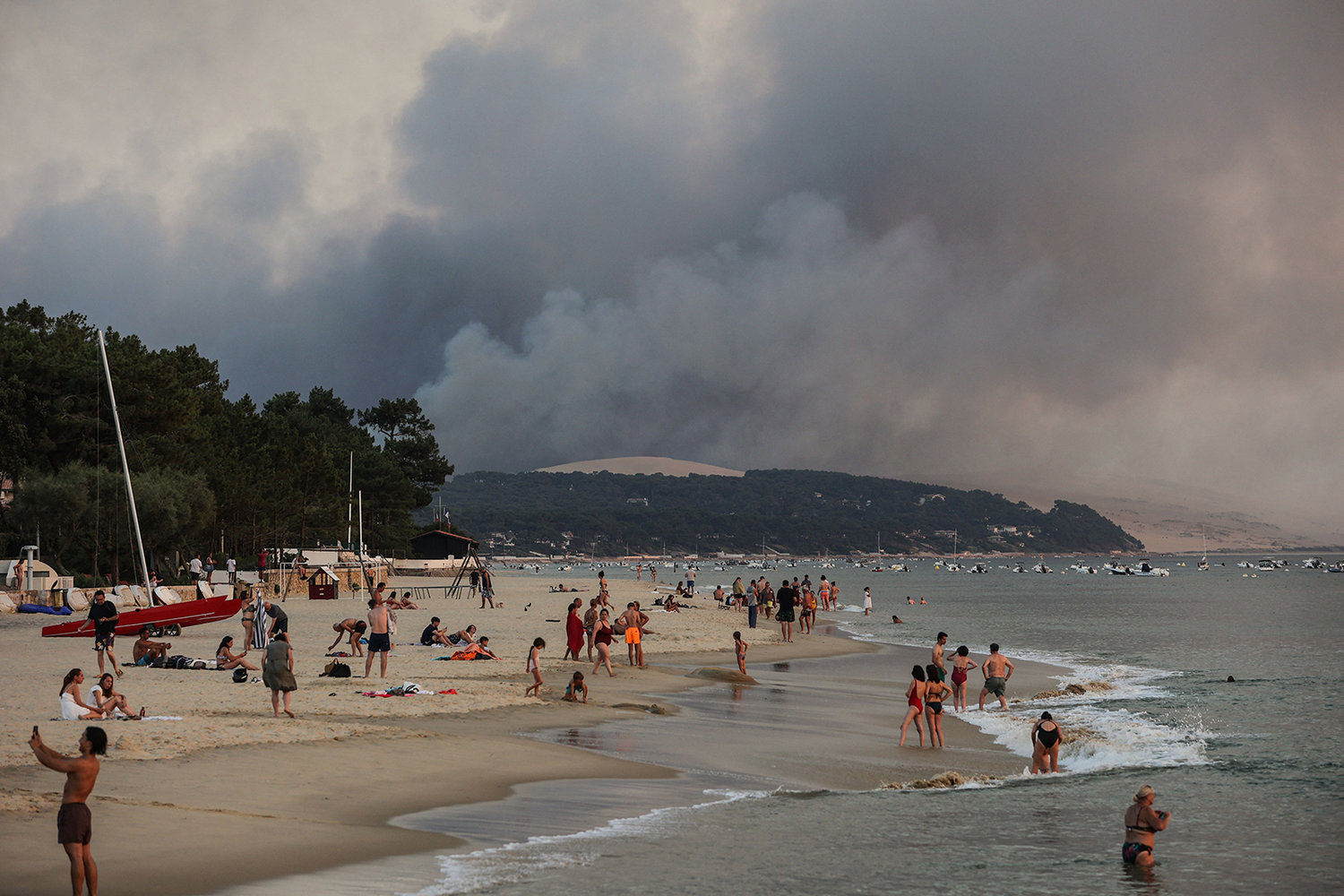 This photograph taken on July 18, 2022, shows people swim on the Moulleau's beach as the smoke rising from the forest fire in La Teste-de-Buch, seen from Arcachon, in front of the Pilat dune. - In scorching heat, with more than 40°C, some 8,000 people had to leave - in a "preventive manner" according to the prefecture - the Miquelots and Pyla-sur-Mer, districts of the municipality of La Teste-de-Buch, a town of 28,000 inhabitants where 4,300 hectares of forest went up in smoke. (Thibaud Moritz/AFP via Getty Images/TNS)