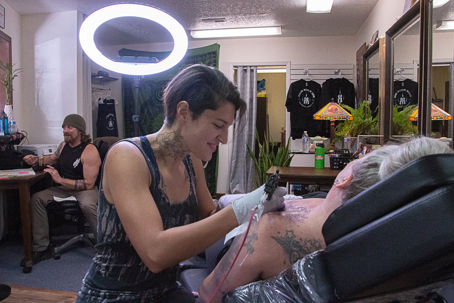 Jen McKay relaxes in the seat while tattoo artist Sarah Gootgeld continues working on a piece on her shoulder while Sarah's husband and receptionist, Eli Gootgeld, enjoys a snack in the back.