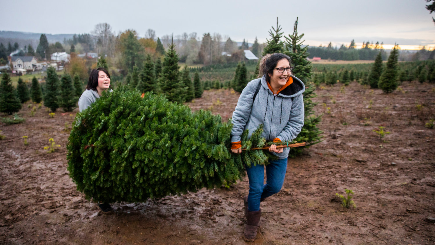 Winona Gladue smiles while carrying a Christmas tree with her daughter Lily, 13, through mud at the Mistletoe Tree Farm west of Chehalis on Friday.