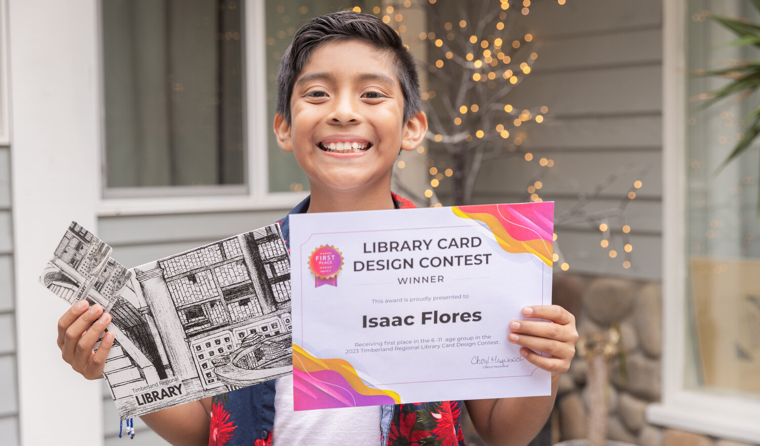 Isaac Flores smiles and holds up a Timberland Regional Library card with a picture of his art on the front alongside a first place certificate for his design at his Centralia residence on Tuesday, Sept. 19.